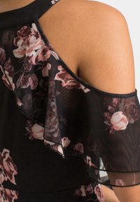 Svart klänning med blommönster, prydd med lager av genomskinliga volanger vid axlarna med rosa och vita rosor. Mjuk textur och figurnära silhuett.