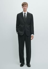 Schwarzer, maßgeschneiderter Anzug, bestehend aus einem einreihigen Sakko mit zwei Taschen und passenden Hosen. Dazu ein weißes Hemd und eine schwarze Krawatte.