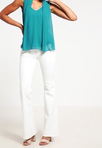 Haut sans manches turquoise avec un décolleté en V et un devant drapé, associé à un pantalon blanc évasé et des sandales à talons carrés beige clair.