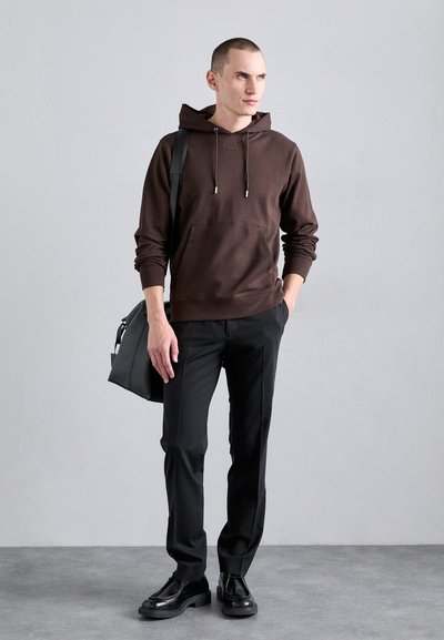 Felpa marrone con cappuccio e coulisse regolabili, tasca frontale, abbinata a pantaloni neri e scarpe nere. Il modello porta una borsa a tracolla grigia.