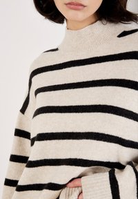 Maglione beige con strisce orizzontali nere, caratterizzato da un collo alto a coste e da un tessuto morbido e testurizzato. Vestibilità ampia con maniche oversize.