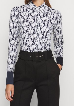 Vrouw draagt een blouse met lange mouwen en een zwart-wit handjespatroon, gecombineerd met zwarte high-waist broek en een brede riem.