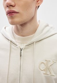 Bézs színű zipzáros kapucnis pulóver texturált "CK" logóval. Cipzáras kapucnis kialakítás, húzózsinóros kapucni és bordázott nyak részlet. Puha anyagú textúra.