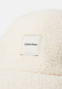 Krémovo sfarbená fleecová čiapka s obdlžnikovou bielou nálepkou s nápisom "Calvin Klein" v čiernej farbe. Mäkká textúra, zakrivený vrch, minimalistický dizajn.