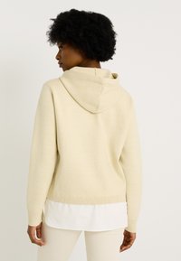 Sudadera beige con capucha y dobladillo acanalado, de textura suave, y una camiseta blanca en capas debajo. El diseño presenta un corte holgado y una costura sencilla.