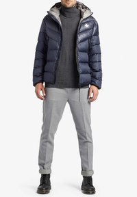 Uomo che indossa un giubbotto imbottito blu navy sopra un dolcevita grigio, pantaloni grigio chiaro con risvolto e stivali neri con lacci, in piedi contro uno sfondo bianco.