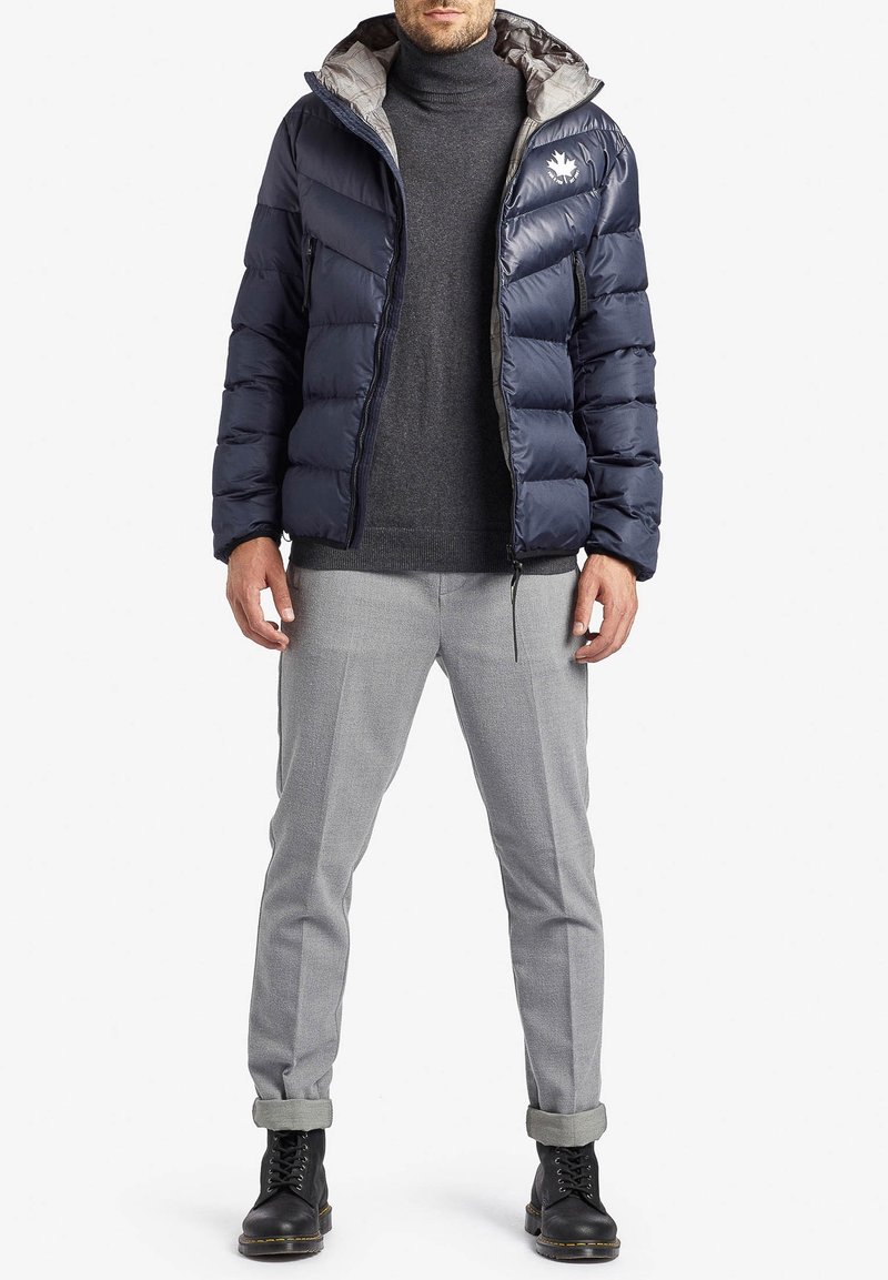 Uomo che indossa un giubbotto imbottito blu navy sopra un dolcevita grigio, pantaloni grigio chiaro con risvolto e stivali neri con lacci, in piedi contro uno sfondo bianco.