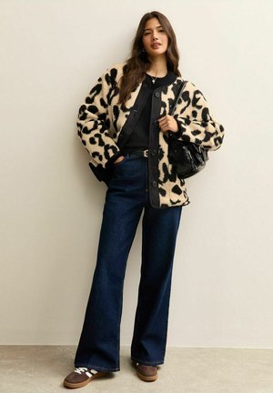 Vrouw die een fleecejack met luipaardprint draagt, een zwarte top, donkere jeans met wijde pijpen, bruine sneakers, en een zwarte handtas vasthoudt.