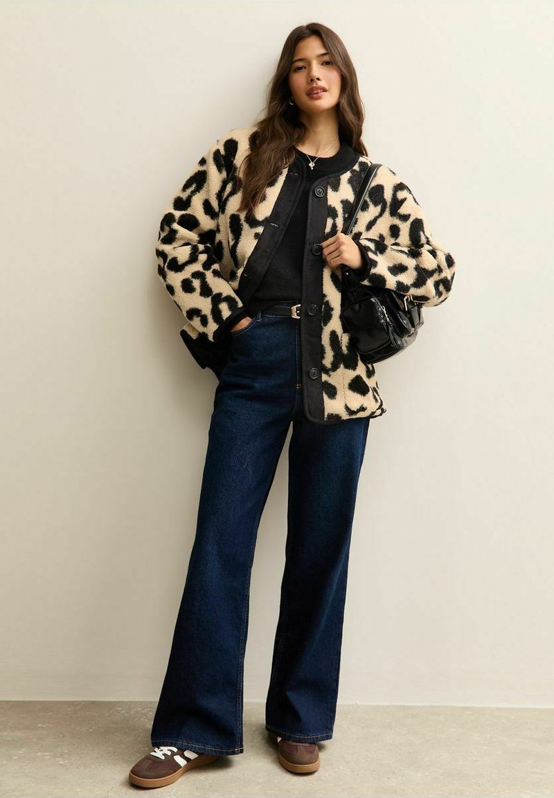Vrouw die een fleecejack met luipaardprint draagt, een zwarte top, donkere jeans met wijde pijpen, bruine sneakers, en een zwarte handtas vasthoudt.