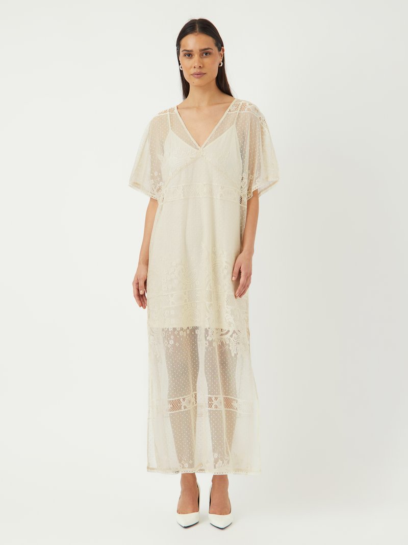 Robe longue crème avec un overlay en dentelle transparente brodée et des motifs à pois. Décolleté en V et manches larges. Associée à des talons blancs.