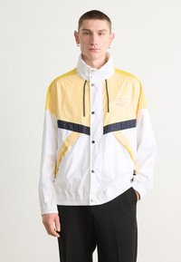 Jachetă de vânt albă și galbenă cu guler ridicat, accente negre, nasturi cu capse și manșete elastice, având un design sportiv și material ușor.