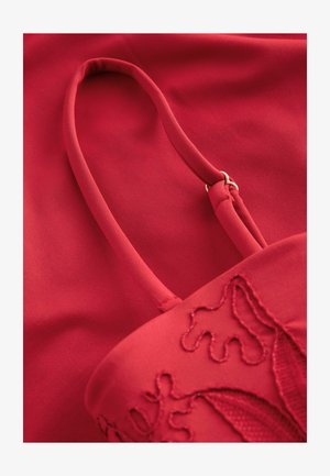 Rdeča tkanina torba z gladko teksturo, ki ima tanko naramnico in zapleteno vezeno cvetlično zasnovo. Celotna barva je globoka, živahna rdeča.