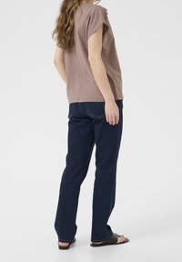 Braunes Kurzarmoberteil mit Schulterdetails, kombiniert mit dunkelblauen geraden Jeans und schwarzen Rutsch-Sandalen.