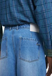 Pantalones cortos de mezclilla azul claro con una textura suave, que cuentan con dos bolsillos traseros y una etiqueta blanca en la cinturilla. Camisa a cuadros horizontales visible.