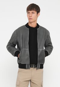 Chaqueta bomber de cuero gris con puños y cuello de canalé, que cuenta con un cierre delantero y dos bolsillos laterales. Llevada sobre una camisa negra.