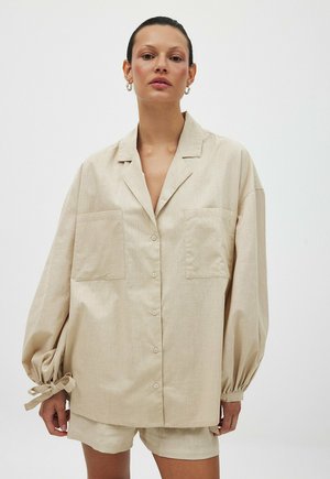 Camicia beige oversize con maniche lunghe e arricciate, due tasche frontali e chiusura con bottoni. Realizzata in tessuto leggero con una finitura testurizzata.