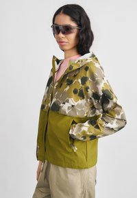 Γυναίκα που φοράει μπουφάν με λουλουδάτο σχέδιο και ελιάς πράσινο, μπεζ παντελόνι και σκουρόχρωμα γυαλιά ηλίου, στέκεται με το ένα χέρι στην τσέπη του μπουφάν.