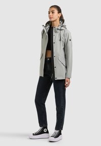 Žena stojaca v svetlosivej kapucňovej bunde cez čierny skrátený top, tmavých džínsoch a čiernych vysokých teniskách na obyčajnom pozadí.