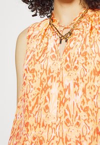Ærmeløs orange bluse med et livligt abstrakt mønster, nøglehulshalsudskæring, samling ved skuldrene og guldkædeaccent.