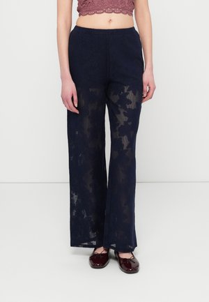 Pantalones de pierna ancha de encaje azul marino con un patrón floral, textura transparente y cintura elástica, combinados con zapatos burdeos brillantes.