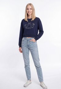 Fiatal, szőke hajú nő tengerészkék, rövid fazonú pulóvert visel, rajta szöveg, világoskék farmert és fehér tornacipőt, egyik kezét a zsebében tartva áll.