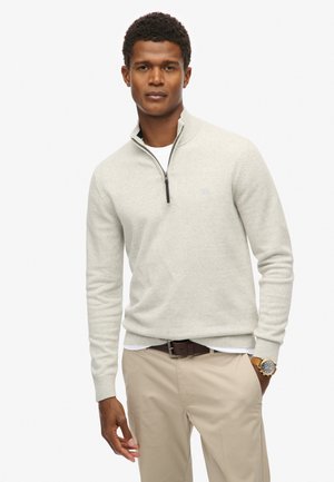 Suéter gris claro con cuello de media cremallera, puños y dobladillo acanalados. Llevado sobre una camisa blanca, combinado con pantalones beige y un cinturón marrón.