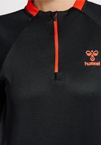 Camiseta atlética negra con un pequeño zipper, acentos rojos en el cuello y un pequeño logo naranja en el lado izquierdo del pecho. Textura suave.