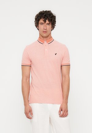 Uomo con capelli ricci che indossa una polo rosa chiaro con colletto e bordi delle maniche a righe, abbinata a pantaloni bianchi, di fronte.