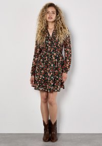 Langärmliges Blumenkleid mit dunklem grünen Hintergrund, das orangefarbene und weiße Blumen zeigt, tailliert in der Taille und mit einem rüschierten Saum. Braune Cowboy-Stiefel.