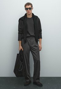Donkergrijs, gestructureerd crewneck sweater onder een zwarte cardigan, gecombineerd met gestreepte broek en zwarte schoenen. Houdt een canvas tote bag vast.