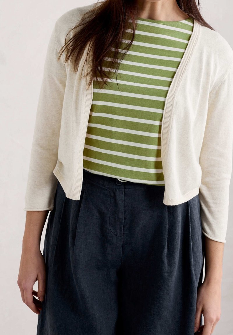 Femme portant une chemise rayée verte et blanche, un cardigan écru raccourci, et un pantalon taille haute foncé, debout devant un fond uni.