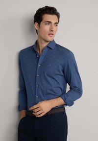 Hombre con camisa azul de botones y mangas arremangadas, pantalones oscuros, de pie frente a un fondo gris claro liso, mirando de lado.