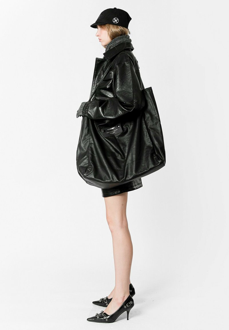 Chaqueta de cuero negra de gran tamaño con acabado texturizado, combinada con una falda corta negra. Los accesorios incluyen una gorra negra y un bolso tote de cuero.