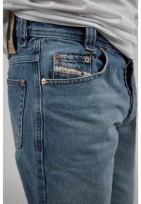 Vaaleansiniset farkut klassisella leikkauksella. Varusteena kuparinväriset yksityiskohdat, takatasku, jossa on logopainatus, sekä hienovaraisesti teksturoitu kangas.