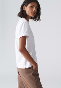 T-shirt blanc à manches courtes avec des poignets retroussés et des fentes sur les côtés, associé à un pantalon marron. Texture lisse et design simple.