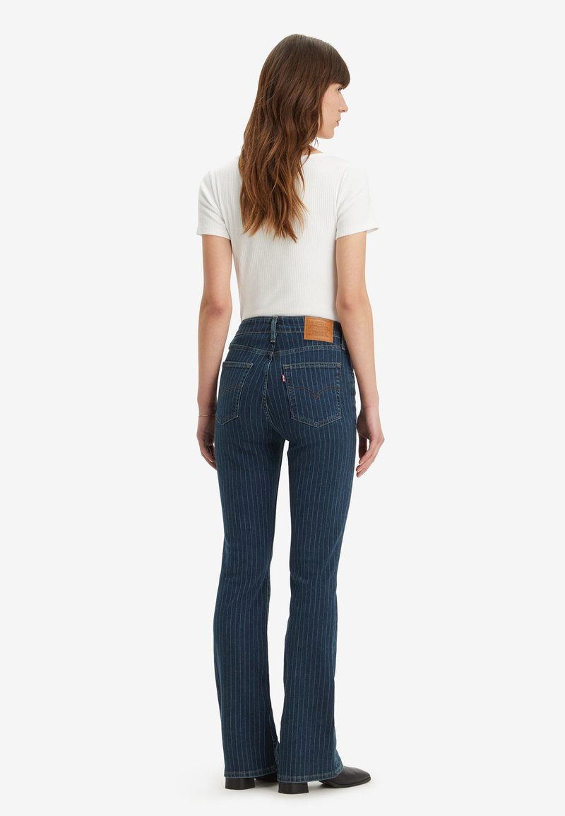 Femme de dos portant un haut blanc à manches courtes en ribbed et un jean flare bleu à rayures avec des chaussures noires sur fond blanc.