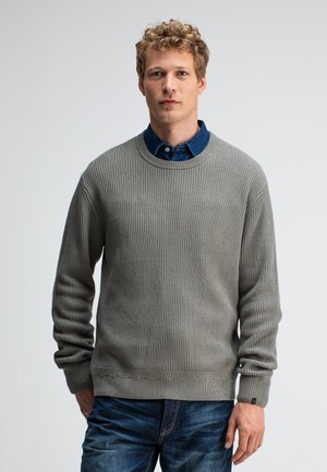 Grijze geribbelde trui met een ronde halslijn en lange mouwen, gecombineerd met een blauwe spijkerhemd eronder, gedragen met een donkere spijkerbroek.