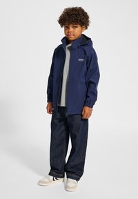 Mörkblå zip-up jacka med huva, elastiska muddar. Bärs över en grå skjorta, ihop med lösa denimjeans och vita sneakers.