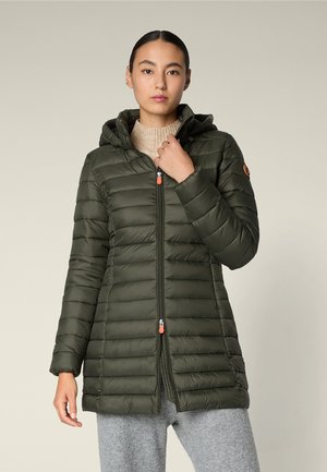 Donna che indossa una lunga giacca trapuntata verde oliva con cappuccio, maglione beige e pantaloni grigi, in piedi davanti a uno sfondo semplice.