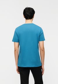 Άνδρας με κοντά μαύρα μαλλιά, φοράει μονόχρωμο μπλε κοντομάνικο μπλουζάκι και μαύρο παντελόνι, στέκεται γυρισμένος με την πλάτη σε λευκό φόντο.