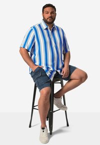 Korte mouwen, blauw-wit gestreept shirt van licht materiaal, gecombineerd met denim shorts en witte sneakers. Zittend op een zwarte barkruk.