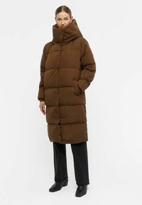 Cappotto imbottito marrone con colletto alto, caratterizzato da un design trapuntato e tasche. Abbinato a pantaloni neri a gamba larga e stivali neri con tacco.