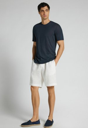 Mann steht mit den Händen in den Taschen, trägt ein marineblaues Kurzarmhemd, weiße knielange Shorts und marineblaue Espadrilles.