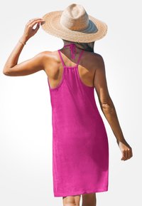 Robe sans manches fuchsia avec un détail de lien dans le dos, texture douce et coupe décontractée. Portée avec un chapeau en paille à large bord.