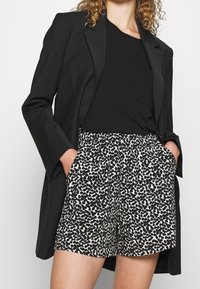Zwarte blazer, zwart T-shirt en korte broek met een wit-zwart abstract patroon, voorzien van zijzakken en een losse pasvorm.