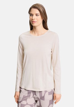 Vrouw met steil bruin haar draagt een beige shirt met lange mouwen en grijze broek met abstracte vormen, staand tegen een effen achtergrond.