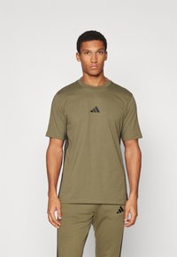 Olive green t-shirt sportowy z krótkim rękawem, okrągłym dekoltem i czarnym logo Adidas na piersi. Gładka tekstura materiału.