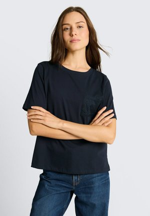 Kobieta z długimi brązowymi włosami, ubrana w czarną koszulkę z krótkim rękawem z teksturowaną kieszenią i niebieskie dżinsy, stoi ze skrzyżowanymi ramionami.