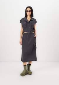 Twee-delig outfit met een cropped, donkergrijze, korte mouwen shirt met zakken en een bijpassende lange, vloeiende rok. Gecombineerd met groene laarzen.