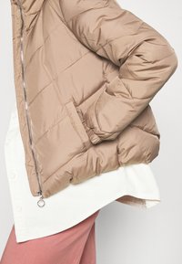 Beżowa kurtka puchowa z zapięciem na zamek, bocznymi kieszeniami i elastycznymi mankietami, nałożona na biały sztruksowy koszula oraz różowe spodnie.